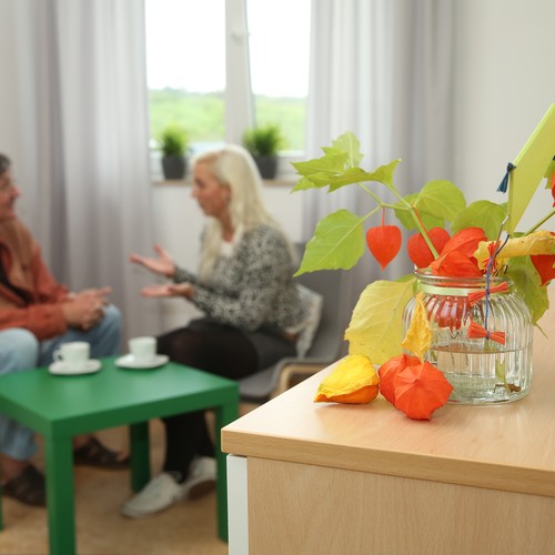 Im Vordergrund steht eine orangefarbene Pflanze, im Hintergrund sieht man einen Mann und eine Frau, die gemeinsam an einem kleinen runden Tisch sitzen