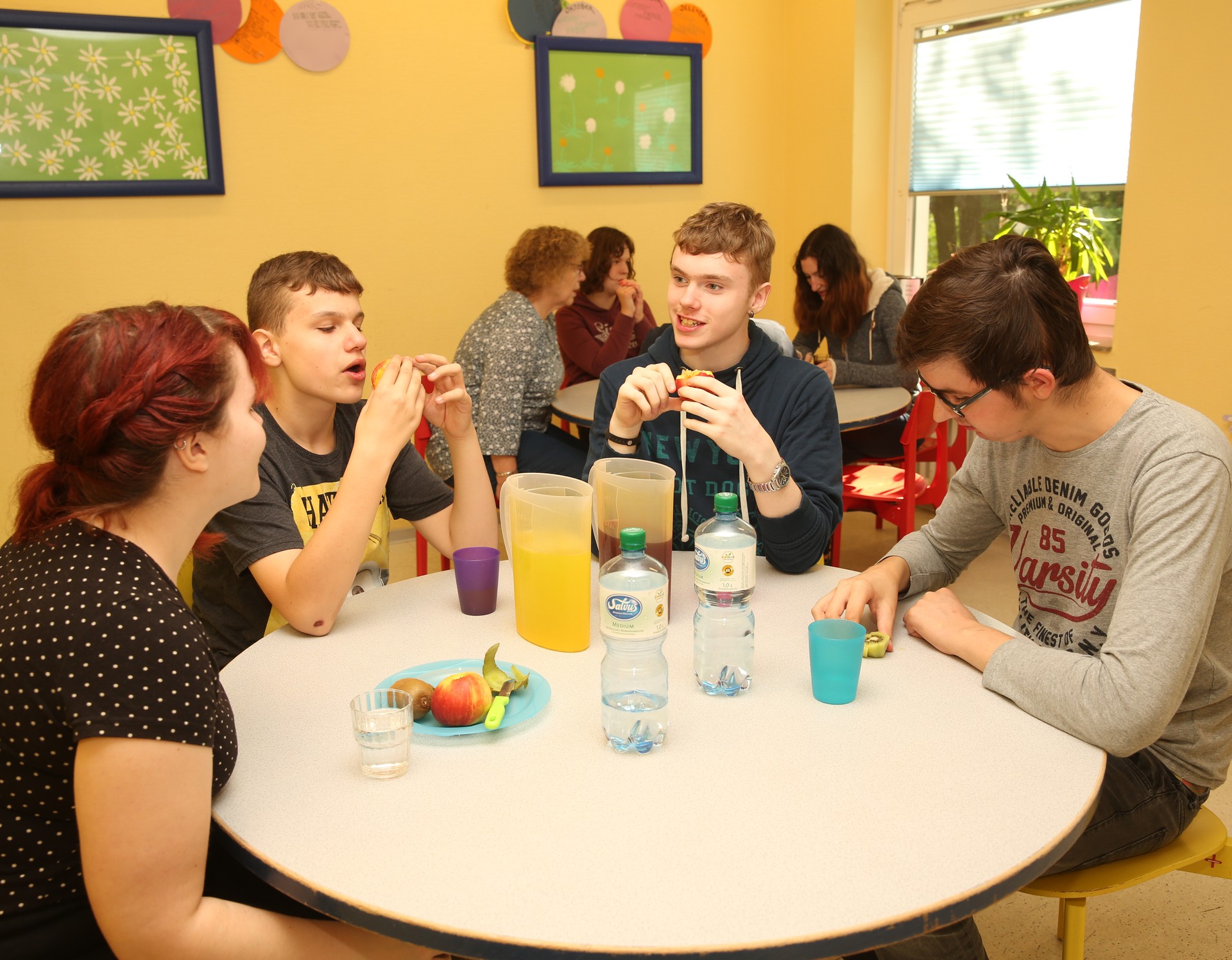 Vier Personen sitzen an einem runden Tisch und essen gemeinsam. Im Hintergrund steht ein weiterer runder Tisch, an dem Menschen gemeinsam essen