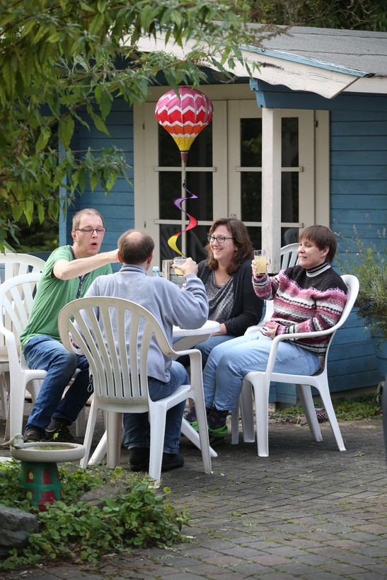 zwei Männer und zwei Frauen sitzen gemeinsam an einem runden, weißen Gartentisch und stoßen mit Gläsern an