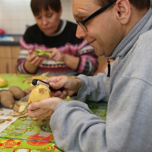 Ein Frau mit pinkem Pullover und ein Mann mit grauem Pullover sitzen gemeinsam an einem runden Tisch und schälen Kartoffeln.