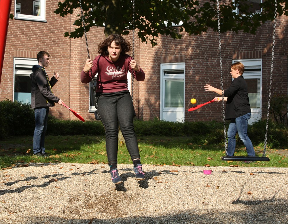 Ein Mädchen in dunkelrotem Pullover schaukelt, im Hintergrund spielen ein Junge und eine Frau Tennis