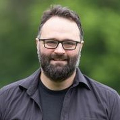 Mann mit dunklem Vollbart und kurzen Harren im karrierten Hemd von einem grünen Hintergrund