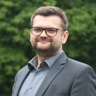 EIn Mann mit kurzen braunen Haaren und Brille, gekleidet mit einem grauen Anzug