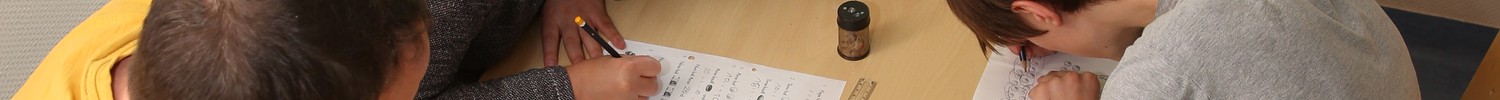 Vier Menschen sitzen gemeinsam an einem eckigen Tisch, jeweils zwei sitzen sich gegenüber. Sie schreiben etwas auf.