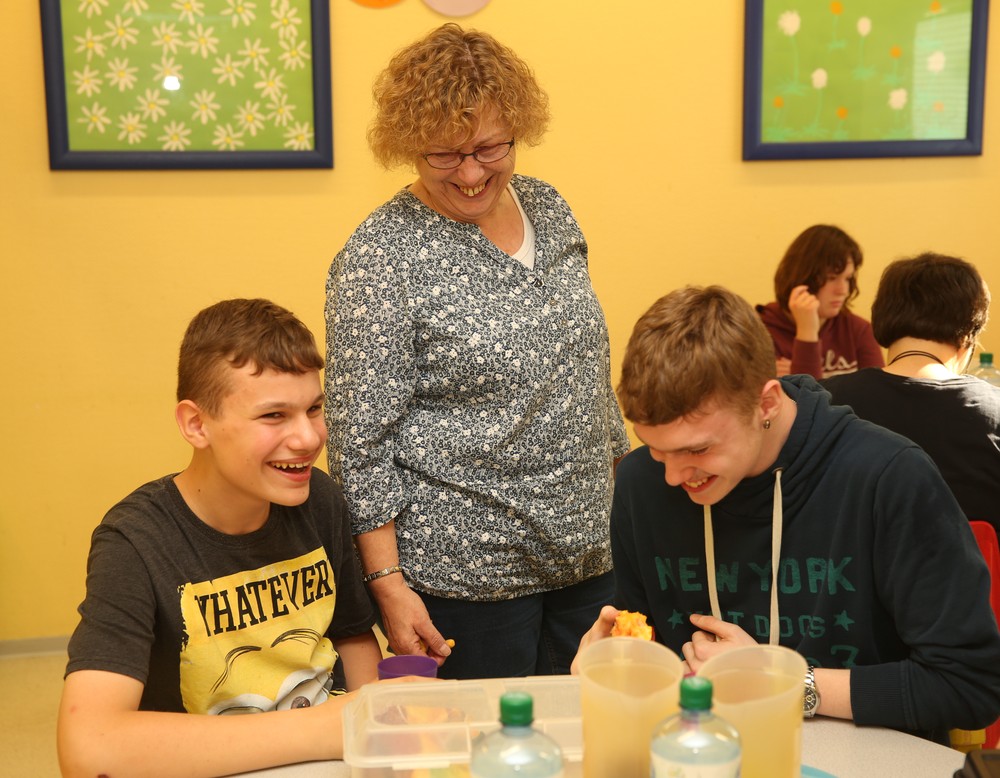 zwei dunkel gekleidete Jungs sitzen zusammen an einem runden Tisch, zwischen ihnen steht eine Frau mit blonden Locken. Alle lachen gemeinsam.