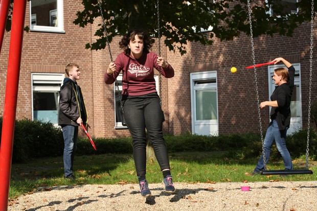 Ein Mädchen mit dunkelrotem Pullover schaukelt. Im Hintergrund spielen ein Junge und eine Frau ein Ballspiel mit Schlägern.