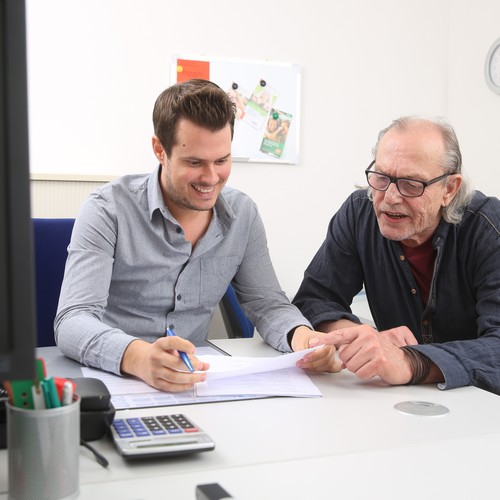 Zwei Männer sitzen nebeneinander, einer hält ein Blatt Papier in der Hand, auf welches beide schauen. Die Männer lächeln.