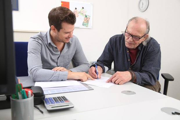 Zwei Männer sitzen zusammen an einem Tisch und schauen gemeinsam auf ein Blatt Papier, der eine Mann füllt dort gerade etwas aus Zwei Männer sitzen zusammen an einem Tisch und schauen gemeinsam auf ein Blatt Papier, der eine Mann füllt dort gerade etwas aus