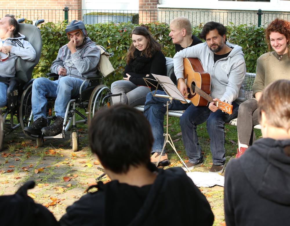 Viele Menschen sitzen zusammen in einem Kreis und musizieren, ein Mann spielt auf einer Gitarre