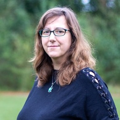 Portrait einer Frau mit langen braunen Haaren, schwarzer Brille und dunkelblauem Pullover vor grünem Hintergrund