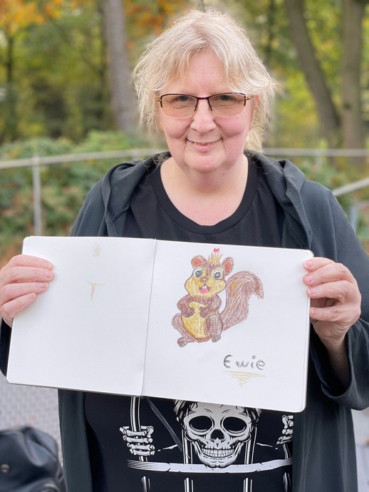 Eine blonde Frau mit Brille lächelt und hält ein Buch (vergrößerte Bildansicht wird geöffnet)