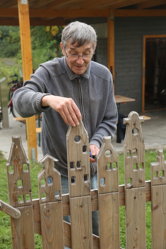 Ein Mann mit grauem Pullover und grauen kurzen Harren steht an einem Holzzaun, dessen Zacken aussehen wie kleine Häuser