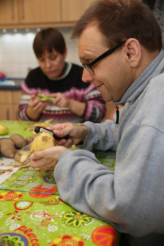 Ein Mann mit kurzen braunen Haaren und Brille sitzt zusammen mit einer Frau mit kurzen braunen Haaren an einem Tisch, sie schälen Kartoffeln