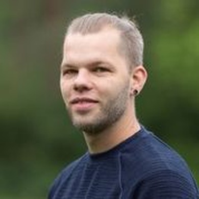 Portrait eines Mannes mit dunkelblonden, zum Zopf gebundenen Haaren und blauem Pullover vor grünem Hintergrund.