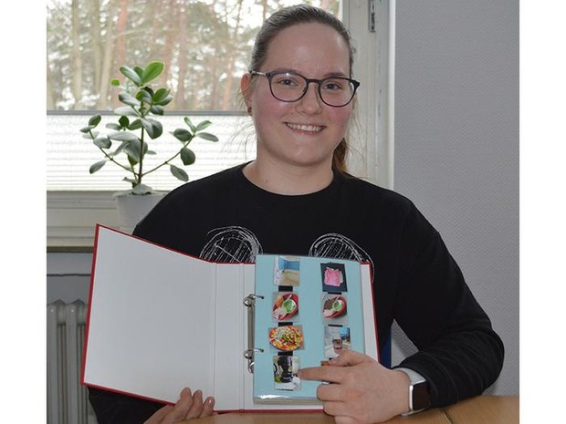 Frau mit Brille und zu einem Zopf gebundenen Haaren hält ein Buch vor sich und lacht