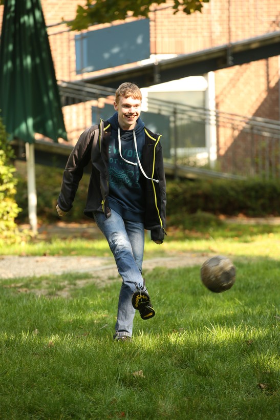Ein Junge mit kurzen Haaren, schwarzer Jacke und blauer Jeans schießt auf einer Wiese einen Ball