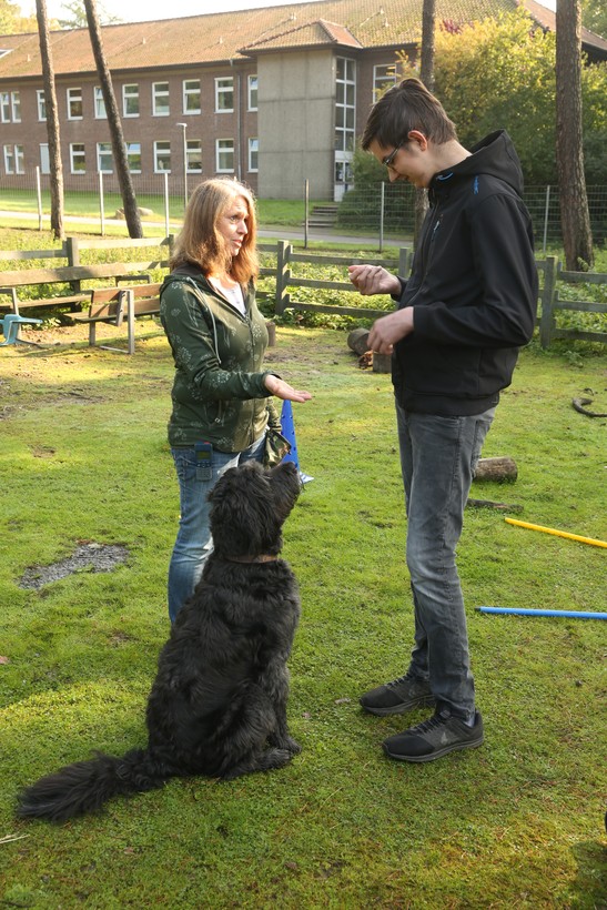 Eine Frau mit grüner Jacke steht zusammen mit einem Jungen mit schwarzer Jacke und einem schwarzen Hund auf einer Wiese. Eine Frau mit grüner Jacke steht zusammen mit einem Jungen mit schwarzer Jacke und einem schwarzen Hund auf einer Wiese.