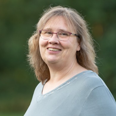 Portrait einer Frau mit dunkelblondem Haar, Brille und grauem Pullover vor grünem Hintergrund