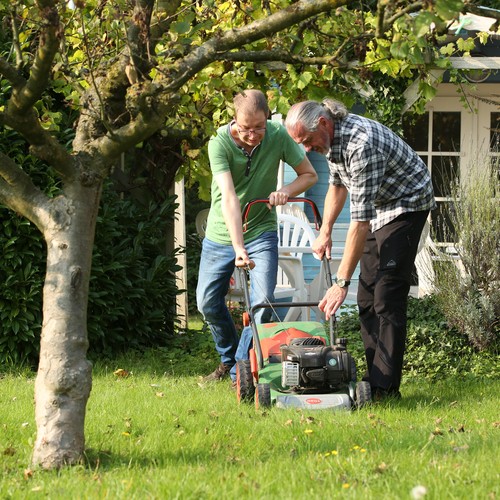 Ein Mann im grünen Shirt mäht den Rasen, ein anderer im karierten Hemd hilft ihm dabei.