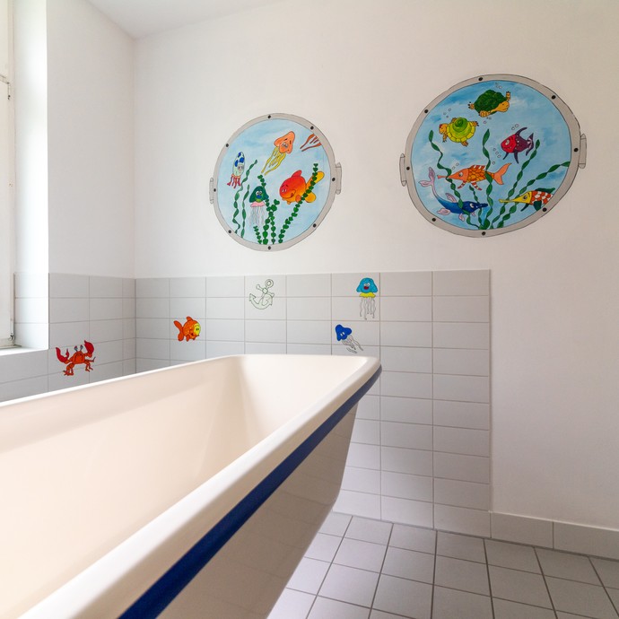 Eine Pflegebadewanne in einem weiß gefliesten Raum, an der Wand sind Fische aufgemalt (öffnet vergrößerte Bildansicht)
