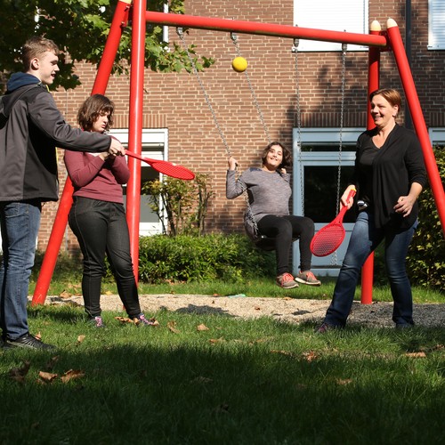 Eine Frau spielt zusammen mit eine Jungen ein tennisähnliches Spiel, im Hintegrund sind zwei Mädchen zu sehen, wovon eine schaukelt.